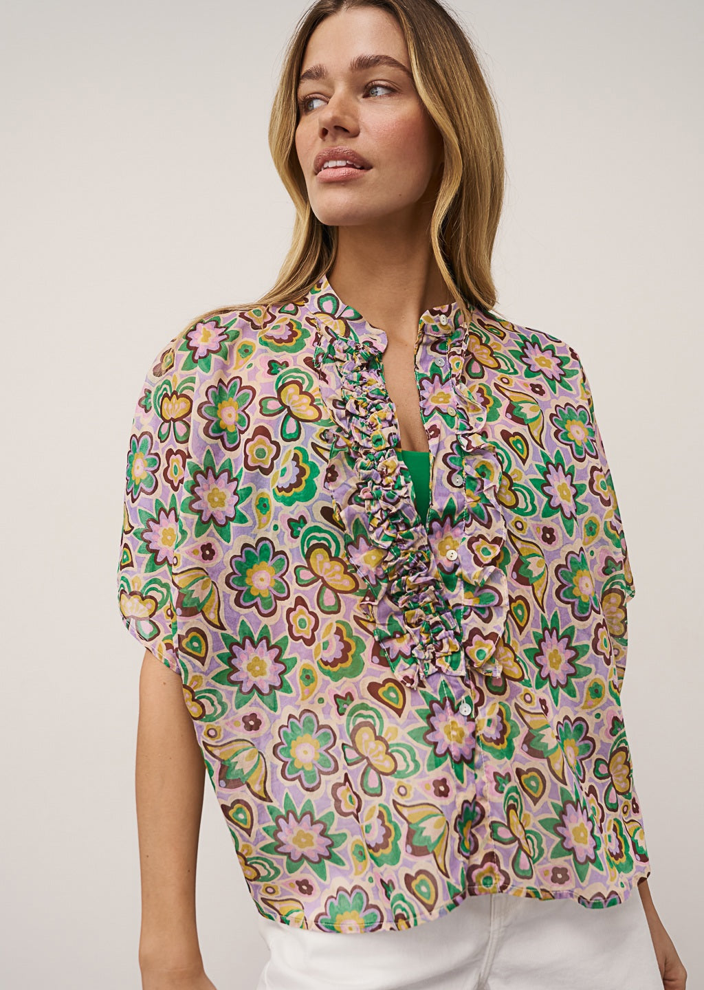 A woman with blonde hair poses casually against a plain background, wearing the 0039 ITALY DAISY BLOUSE IN LILAC AND GREEN PRINT, styled with white shorts and beige flats.