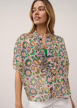 A woman with blonde hair poses casually against a plain background, wearing the 0039 ITALY DAISY BLOUSE IN LILAC AND GREEN PRINT, styled with white shorts and beige flats.
