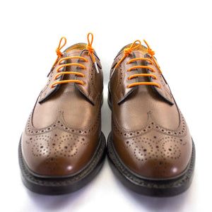 A pair of brown leather brogue shoes with decorative perforations features STOLEN RICHES - DRESS SHOELACE IN TIQUI ORANGE by STOLEN RICHES, shown on a white background.