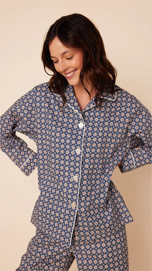 A person stands against a beige background wearing the CAT'S PAJAMAS Newport Daisy Luxe Pima Cotton Pajamas by CAT'S PAJAMAS. The navy blue pajama top features a white geometric pattern, long sleeves, a collar, and white buttons down the front. The matching full-length pajama pants complete the set. The person is barefoot.
