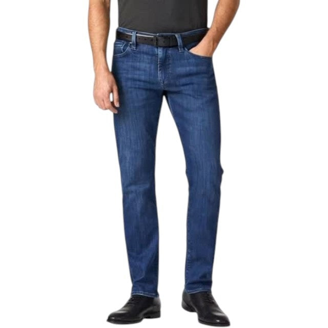 A man stands facing forward, wearing a dark short-sleeve polo, 34 HERITAGE - COURAGE STRAIGHT LEG JEAN IN MID URBAN WASH, a black belt, and black shoes. One hand is in his pocket; he has short hair, a trimmed beard, and a neutral expression.