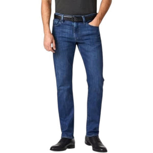 A man stands facing forward, wearing a dark short-sleeve polo, 34 HERITAGE - COURAGE STRAIGHT LEG JEAN IN MID URBAN WASH, a black belt, and black shoes. One hand is in his pocket; he has short hair, a trimmed beard, and a neutral expression.