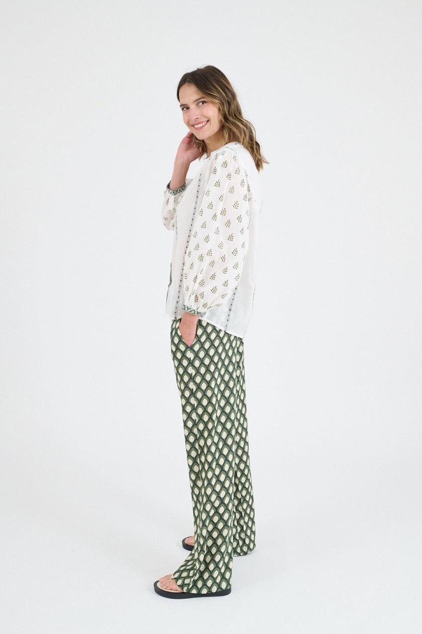 A woman wears the STORIATIPIC LUCIE FLOWER BLOUSE—a cotton blouse with patterned sleeves and green trim—paired with wide-leg green patterned pants, standing against a white background in sandals, evoking casual elegance.