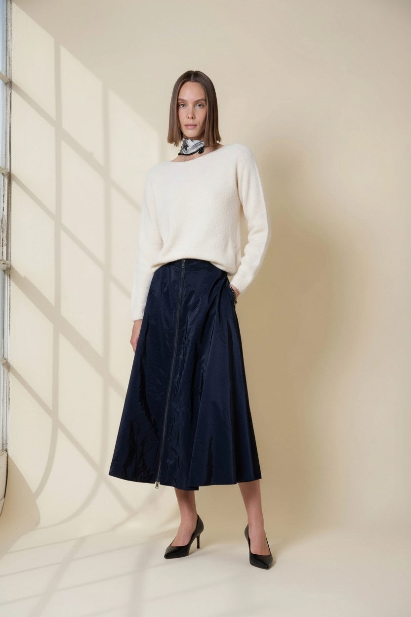 A woman in a cream sweater, scarf, and black heels stands by a large window, sunlight casting shadows on a beige wall, wearing the VICARIO CINQUE gazelle midi taffeta skirt with zipper in midnight blue.
