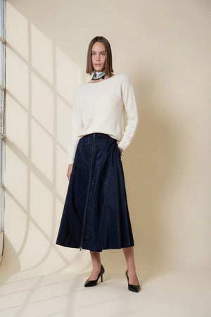 A woman in a cream sweater, scarf, and black heels stands by a large window, sunlight casting shadows on a beige wall, wearing the VICARIO CINQUE gazelle midi taffeta skirt with zipper in midnight blue.