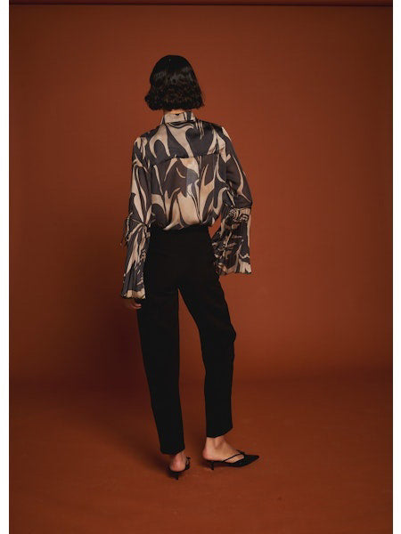 A woman poses against a brown background wearing the MILVA MI black and gold flare sleeve blouse by MILVA, paired with high-waisted black pants. Her dark hair is styled in a low bun.