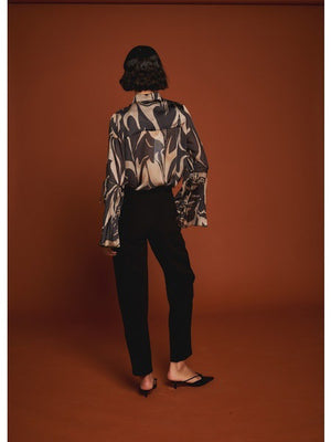 A woman poses against a brown background wearing the MILVA MI black and gold flare sleeve blouse by MILVA, paired with high-waisted black pants. Her dark hair is styled in a low bun.