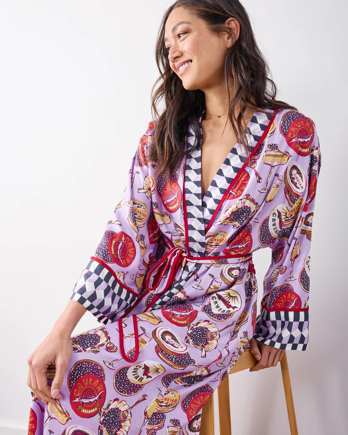 A woman stands barefoot against a white wall, smiling softly in the PRINTFRESH - ECO SATIN ROBE IN CHAMPAGNE AND CAVIAR by PRINTFRESH, featuring colorful food and champagne illustrations, checkered trim, and a matching waist tie.