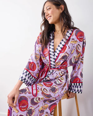 A woman stands barefoot against a white wall, smiling softly in the PRINTFRESH - ECO SATIN ROBE IN CHAMPAGNE AND CAVIAR by PRINTFRESH, featuring colorful food and champagne illustrations, checkered trim, and a matching waist tie.