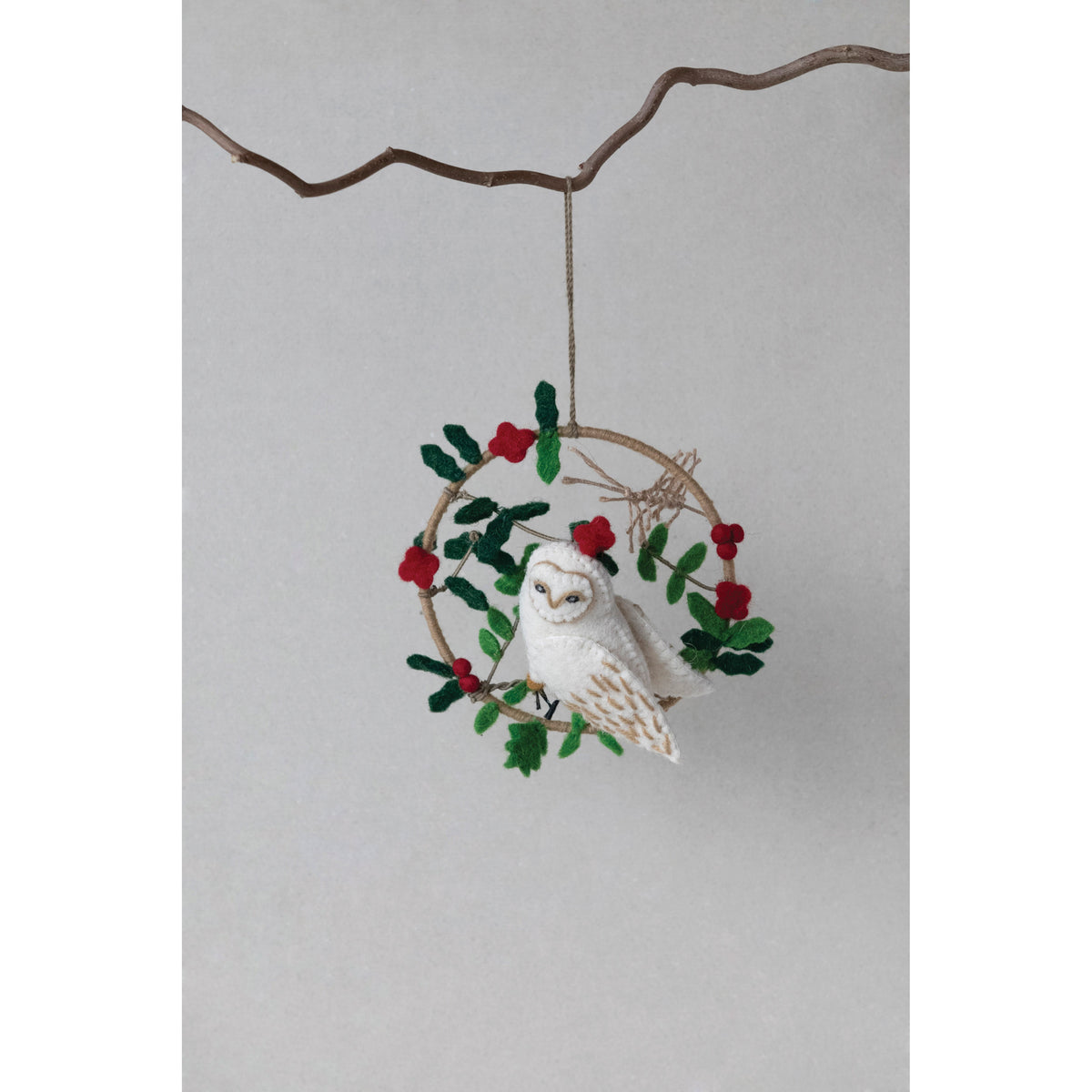 The CREATIVE COOP FELT OWL IN WREATH is a handmade ornament featuring a white felt owl on a circular wreath with green leaves and red berries, hanging by a string loop.