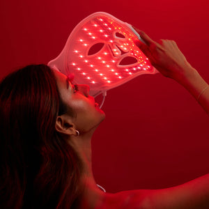 The OMNILUX - CONTOUR FACE MASK by OMNILUX, designed for skin rejuvenation and anti-aging, features red light therapy technology. It has adjustable straps, cutouts for eyes, nose, mouth, and a geometric green-white pattern on a plain white background.