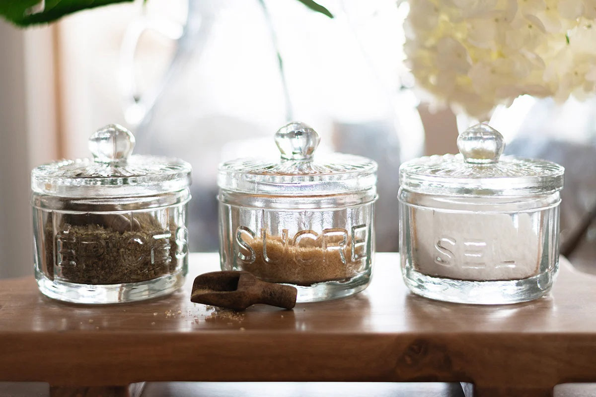 The ETUHOME Bistro Glass Sugar Cellar with Scoop by ETUHOME holds brown sugar on a white countertop near a folded white towel with red stripes and a wooden tray, adding classic charm to your kitchen storage collection.