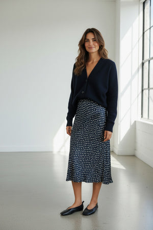 A woman stands in a bright room wearing the HARTFORD - JIMO Bias Cut Viscose Skirt in Navy with White Dot, paired with a navy cardigan and black flats. She has wavy brown hair and is smiling softly as natural light streams through large windows.