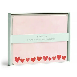 The E. FRANCES - FLAT NOTES BLUSH MINI HEARTS by E. FRANCES PAPER are pale pink notecards with rounded corners and red and pink hearts along the bottom, stacked atop light gray envelopes.