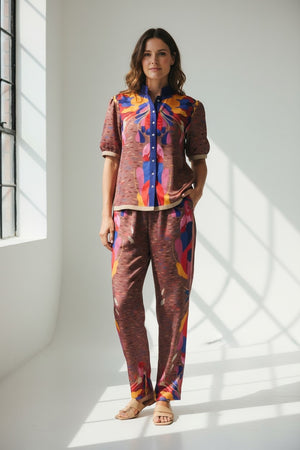 A woman stands indoors in natural light, wearing the FRANCO FERRARI Cotton Print Short Sleeve Blouse with matching loose pants. The set features a statement print in brown, blue, yellow, and pink. She completes her look with sandals.