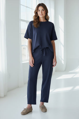 A woman with wavy brown hair smiles as she stands in a bright room, wearing the FRANK & EILEEN Bibi Barrel Leg Sweatpant in Classic Navy paired with a matching short-sleeve top and tan loafers.