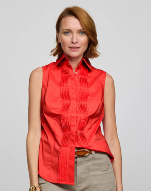 A person with short red hair smiles in a tailored HINSON WU Lilith Sleeveless Shirt featuring decorative pleats, paired with a brown belt and khaki pants against a plain light gray background.