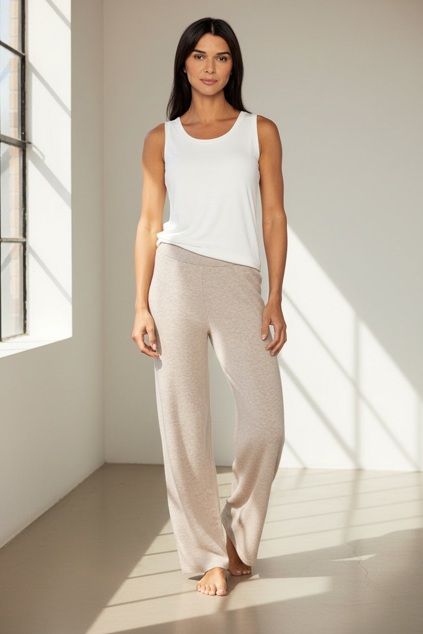 A woman stands against a plain white background, wearing loose light beige pants—the KINROSS CASHMERE Doubleknit Cotton Cashmere Pant by KINROSS—paired with a sleeveless white top and white sneakers. With long dark hair and a relaxed expression, her natural stance highlights these softly tailored pants as an understated addition to any elegant wardrobe.