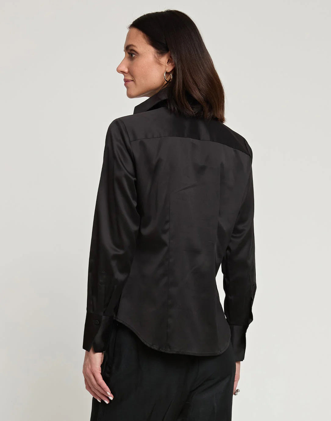 A woman with shoulder-length dark hair is shown from the back, wearing a white HINSON WU - MAYA Long Sleeve Cotton Satin Shirt and black pants. She is standing against a plain, light-colored background.