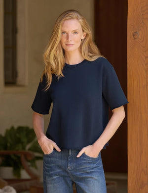 A woman with long blonde hair, dressed in the FRANK & EILEEN Perry Sweatshirt Tee in British Royal Navy and blue jeans, stands indoors and glances over her shoulder at the camera.