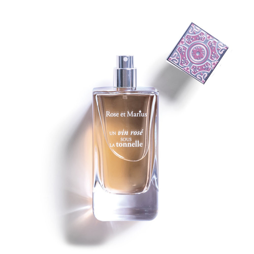 A clear glass bottle of ROSE ET MARIUS - A ROSE WINE UNDER THE ARBOR Eau de Parfum 100ml, known for its fruity woody scent, with a patterned square cap beside it on a white background. The bottle casts a soft shadow.