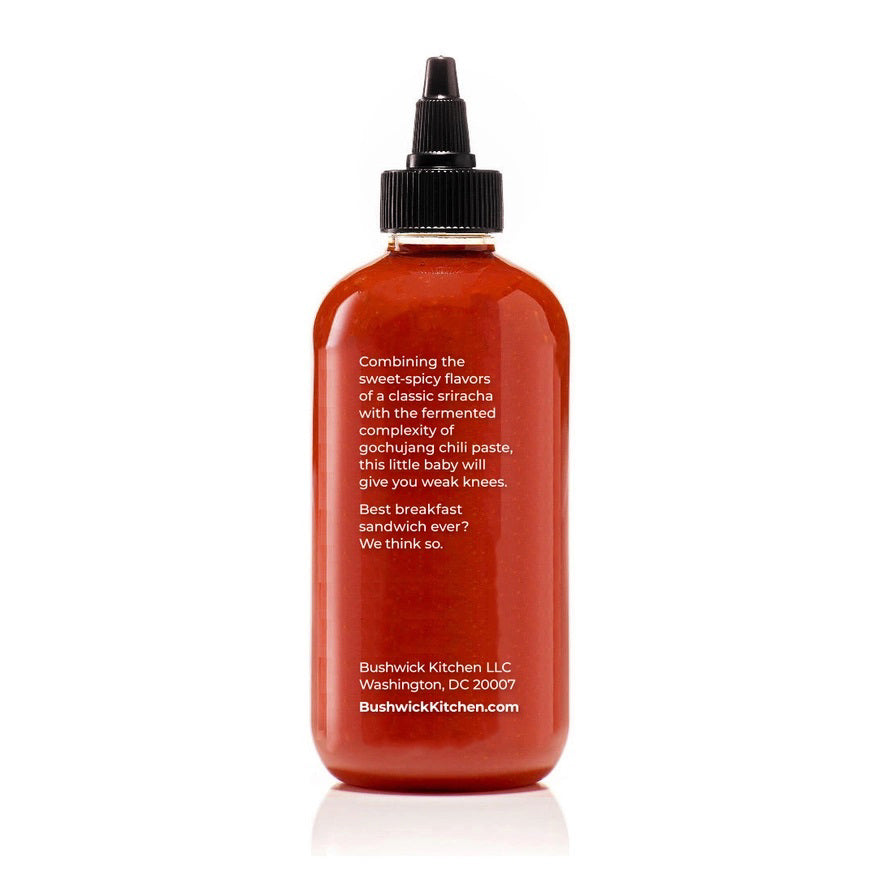 A 10.5 oz (298g) bottle of BUSHWICK KITCHEN - 'WEAK KNEES' GOCHUJANG SRIRACHA hot sauce features a striking red design with a black cap and white text, proudly made in Brooklyn, NY, USA. This unique blend includes fermented chile paste for an exceptional kick.