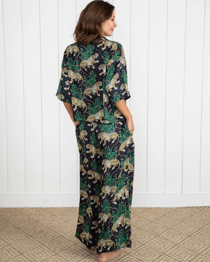 A woman stands barefoot on a woven rug in front of a white paneled wall, wearing the PRINTFRESH - Bagheera Satin Wildest Dream Pajama Set in Ink from PRINTFRESH, featuring a vibrant leopard and foliage pattern with wide-leg pants.