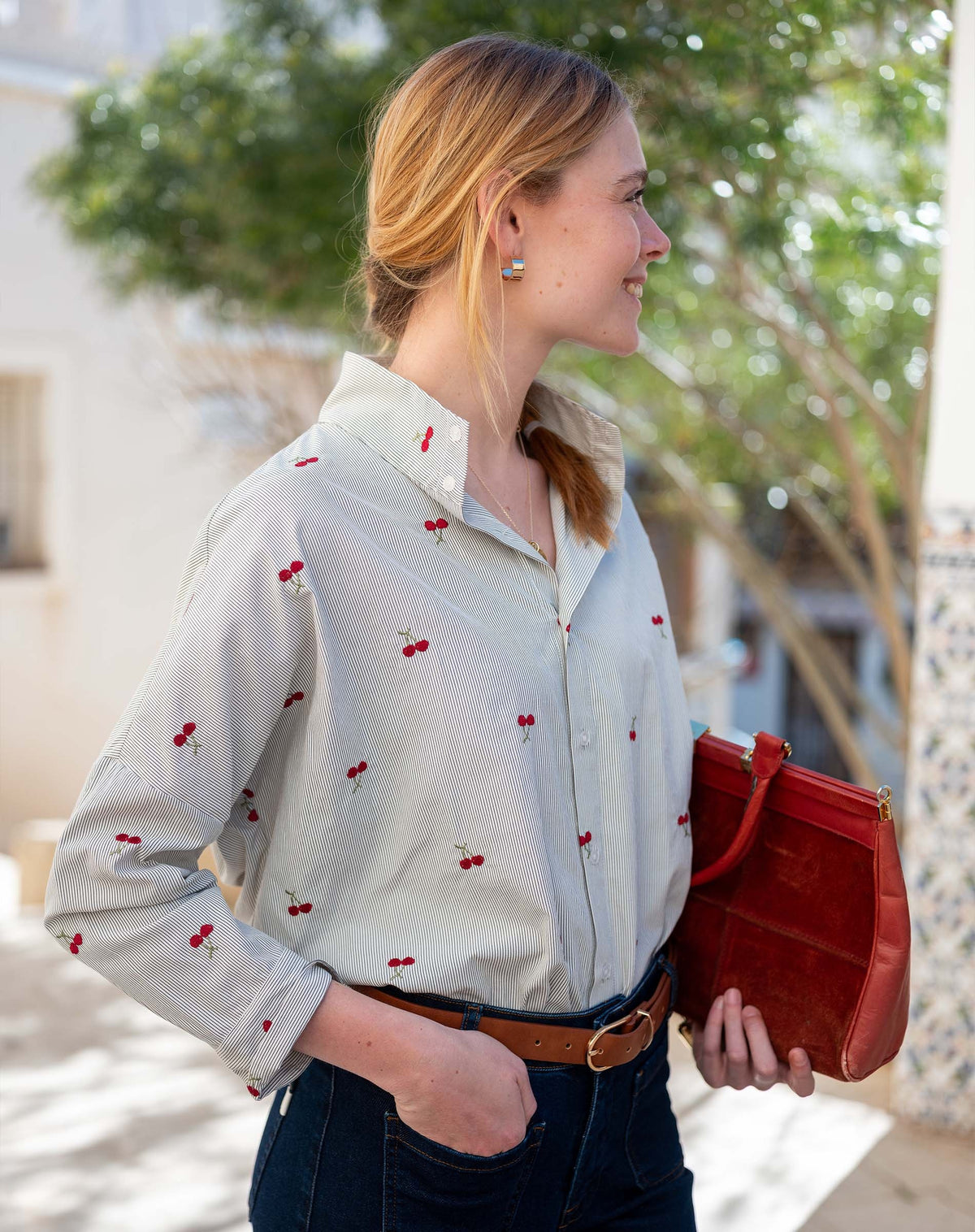 A woman with blonde hair in a ponytail, dressed in the MERSEA Emerson Button Up Shirt in Cherries On Top by MERSEA and blue jeans, holds a red clutch while standing outdoors near green trees and patterned tiles, smiling and looking to the side.