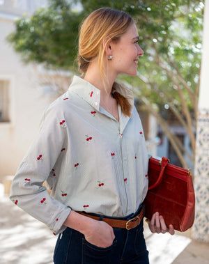 A woman with blonde hair in a ponytail, dressed in the MERSEA Emerson Button Up Shirt in Cherries On Top by MERSEA and blue jeans, holds a red clutch while standing outdoors near green trees and patterned tiles, smiling and looking to the side.