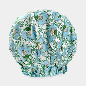 The BATH ACCESSORIES COMPANY offers the BOUFFANT SHOWER CAP, featuring a blue floral and green leaf pattern on a light base. It includes waterproof inner-layers and gathered edges for a puffy, rounded shape.