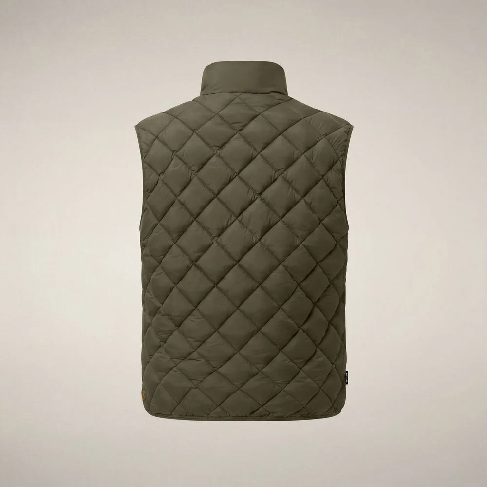 A person stands facing forward, wearing the SAVE THE DUCK - FABRICE Puffer Vest in Snake Green with Plumtech® padding over a beige sweater, paired with white pants and tan shoes, against a plain light background.