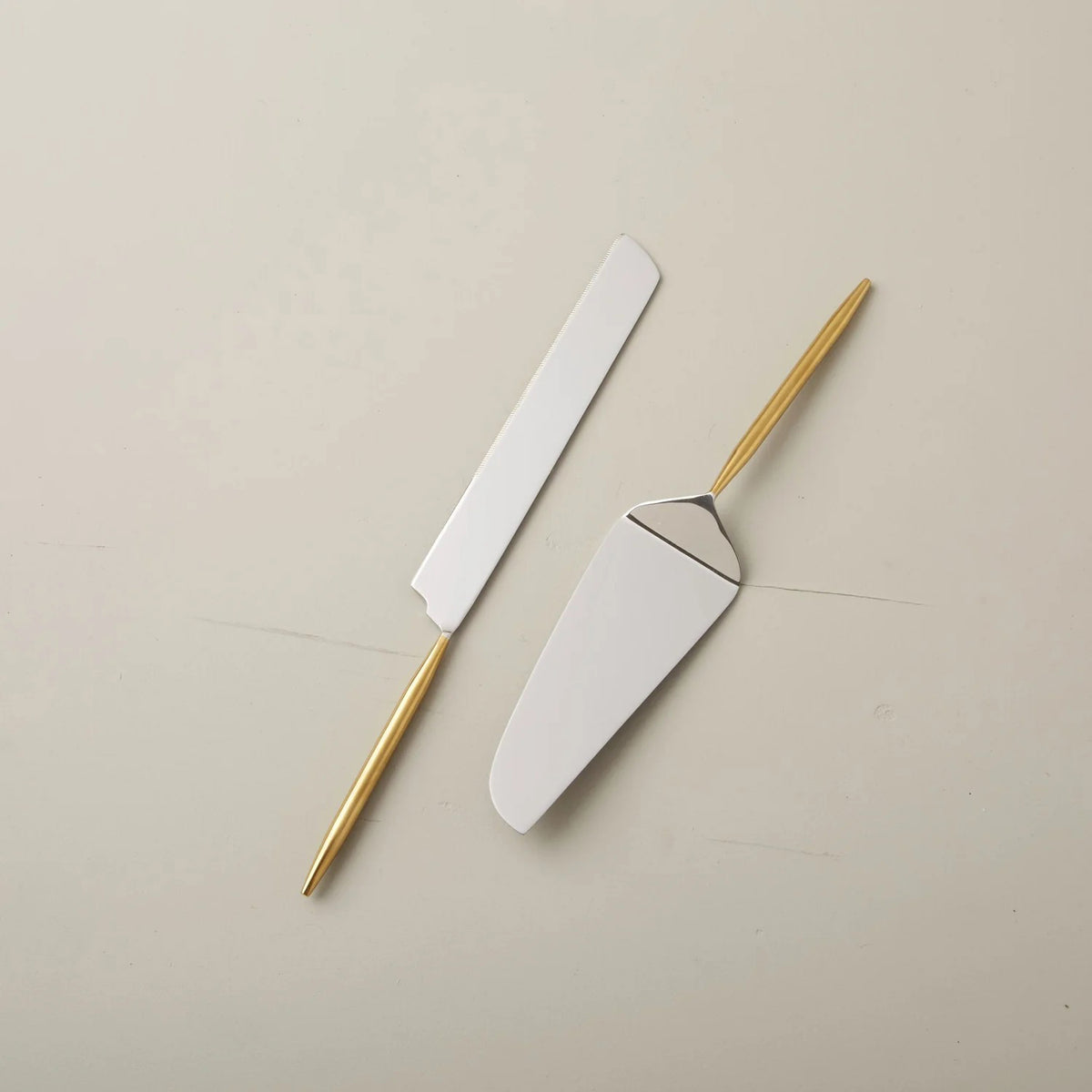 The SUN &amp; MOON CAKE LIFT SET by BE HOME, INC is elegantly displayed diagonally on a plain, light beige surface. The set includes a pie server with a wide, triangular blade and a cake knife featuring a long, narrow blade. Both pieces showcase a sleek, modern design and are handcrafted with a durable PVD finish.