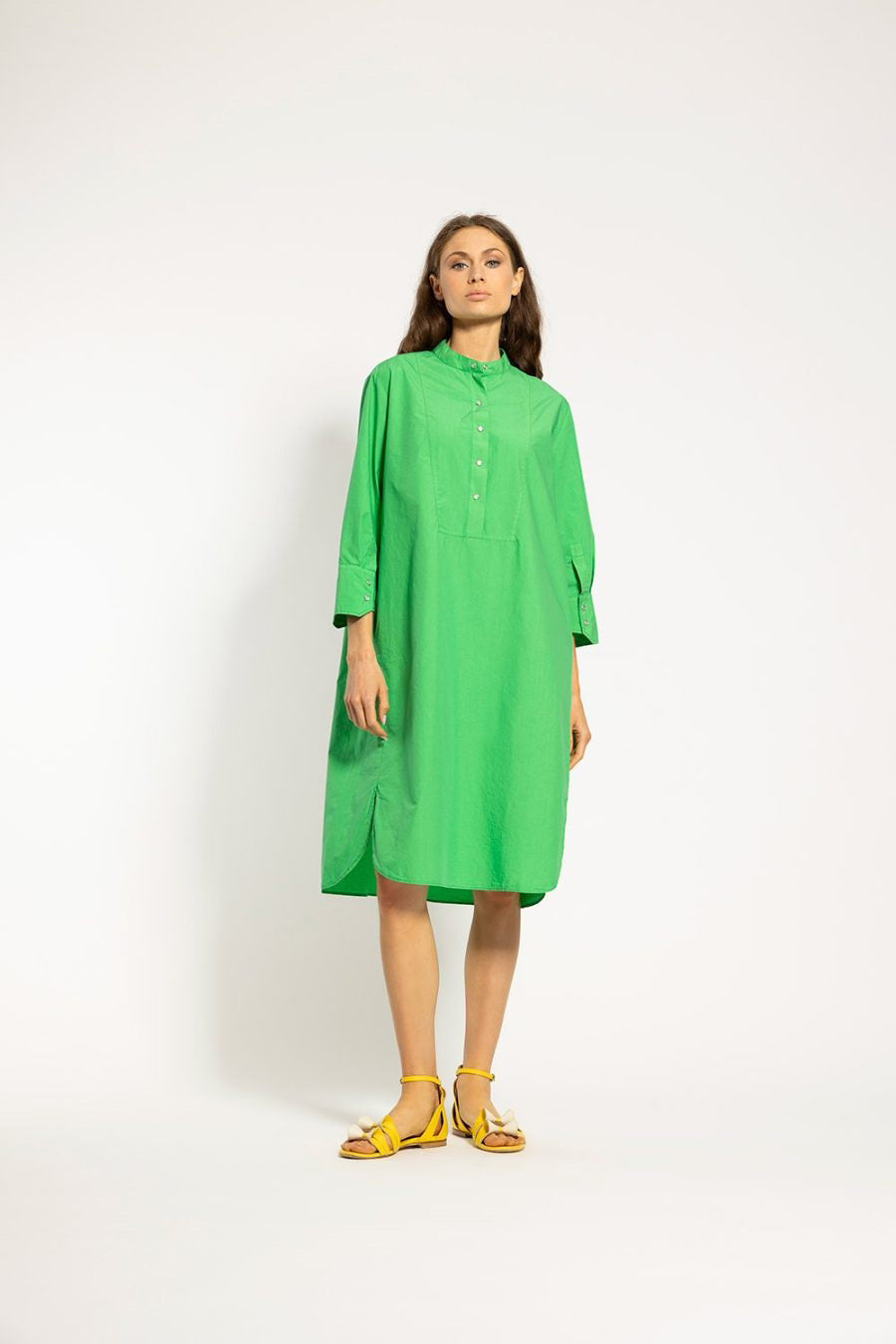 A woman stands against a plain background wearing the ACADEMIA Claude Shirt Dress/Long Tunic in a loose-fitting, bright green style with long sleeves and buttons down the front. She has her hands in her pockets and is looking to the side. Made from 100% cotton by ACADEMIA, she pairs the dress with yellow strappy sandals.