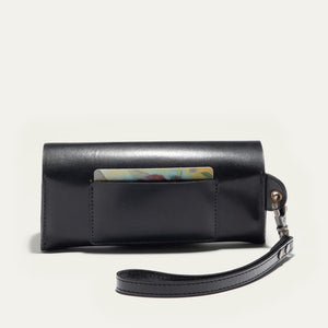 The WILL LEATHER GOODS - EYEGLASS CASE IN BLACK is a handcrafted Italian leather case with a flap closure, embossed logo on the front, and an attached wrist strap on the left, shown against a plain light background.