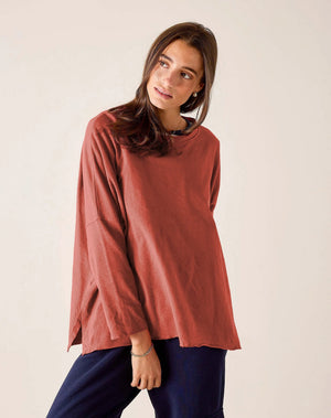 A woman with long brown hair wears the MERSEA Catalina Rustic Tee in Cayenne, paired with dark blue pants. She stands against a plain light background, resting her chin on her hand and gazing at the camera.