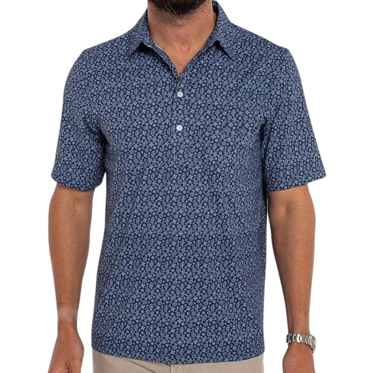 A man wears the CRIQUET Performance Range Polo Shirt in Bandana Navy—a short-sleeve, button-up polo with a white geometric pattern—paired with beige pants and a watch. He is facing away, so his face isn’t visible.