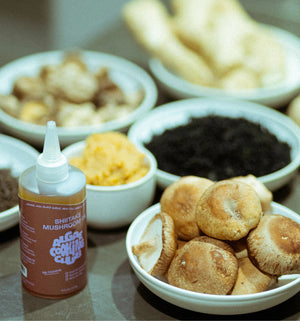 A bottle of ALGAE COOKING CLUB - SHIITAKE MUSHROOM OIL sits on a counter, surrounded by bowls of fresh shiitake mushrooms, dried ingredients, and assorted foods.