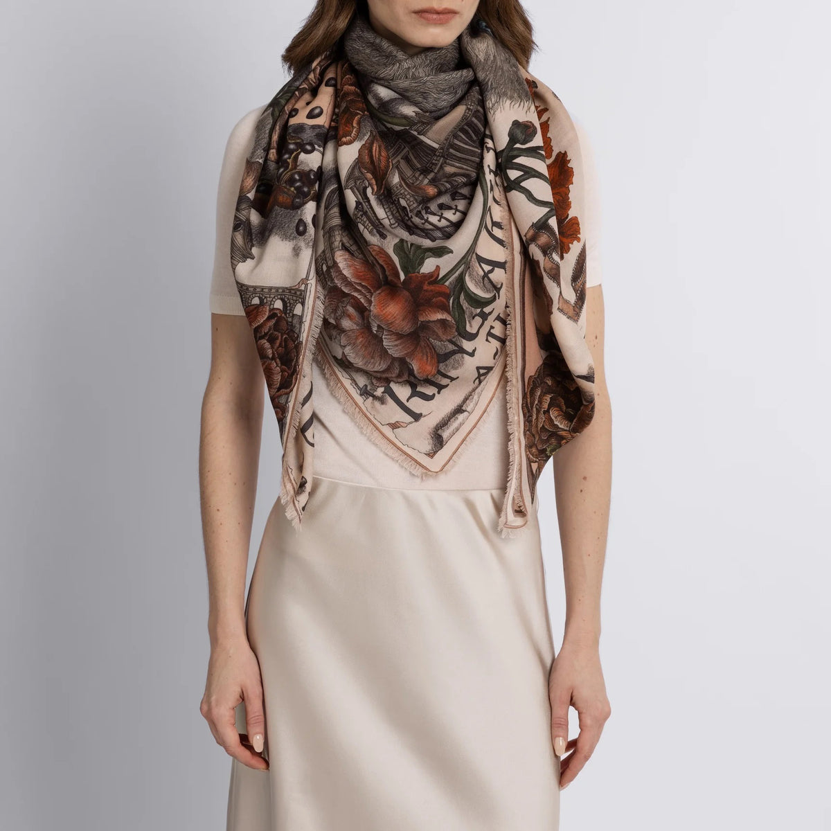 A woman in a beige dress is draped in the Sabina Savage "Ring a Ring o' Roses" silk twill scarf, featuring intricate English garden flower patterns on a marshmallow/ballet background, adding elegance to her ensemble.