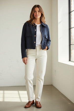 A woman with long brown hair wearing the FRANK & EILEEN Rowan Cropped Sweatshirt Utility Jacket in Triple Terry Classic Navy, paired with white pants, stands sideways and smiles outside near a white building with windows.