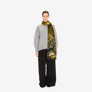 A woman in a light gray sweater and black pants stands against a plain white background, wearing the INOUI EDITIONS - PAPILLON (BUTTERFLIES) wool scarf in emerald from INOUI, featuring black and yellow butterfly designs.