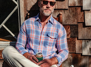 Close-up of a person's chest in the CRIQUET - DOUBLE CLOTH FLANNEL PEARL SNAP SHIRT IN SUNSET, a blue, white, and orange plaid flannel with pearl snaps, front pocket with flap, and visible chest hair at the open collar.