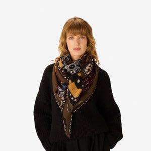 The INOUI EDITIONS - PAPILLONS WOOL SCARF IN KHAKI by INOUI is a square scarf made in India, featuring an intricate and colorful butterfly pattern in brown, blue, yellow, and white hues on a tan background. Measuring 130 x 130 cm, it provides generous coverage and stylish versatility for any occasion.