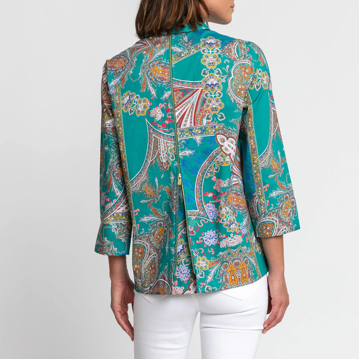The individual is wearing the Hinson Wu Xena Shirt, a teal paisley print blouse with 3/4 sleeves and a collar. The blouse showcases an intricate design featuring vibrant colors like orange, purple, and blue. They have paired it with white pants against a plain white background.