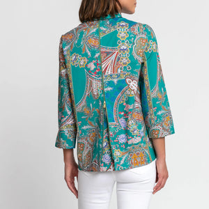 The individual is wearing the Hinson Wu Xena Shirt, a teal paisley print blouse with 3/4 sleeves and a collar. The blouse showcases an intricate design featuring vibrant colors like orange, purple, and blue. They have paired it with white pants against a plain white background.