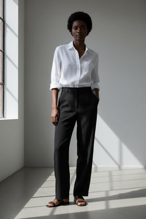 A person with curly hair wears the FRANK & EILEEN EILEEN Relaxed Button Up in Washed Linen White, standing outdoors against a blurred background of greenery and a building.