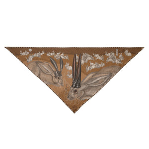 A person dressed in a light beige outfit faces away from the camera, showcasing the SABINA SAVAGE ENGLAND - THE FLOATING RABBITS TRIANGLE SILK TWILL SCARF, a 100% silk twill accessory adorned with whimsical black-tailed jackrabbits wearing top hats. The backdrop is a plain light gray.