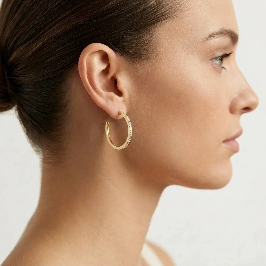 A woman with brown hair in a low bun is shown in profile against a plain light background, wearing the ELLA STEIN - A HOOP THING EARRINGS by ELLA STEIN—gold-plated hoops encrusted with small stones.