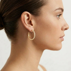 A woman with brown hair in a low bun is shown in profile against a plain light background, wearing the ELLA STEIN - A HOOP THING EARRINGS by ELLA STEIN—gold-plated hoops encrusted with small stones.