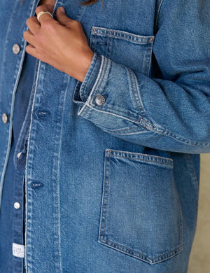 A woman stands indoors wearing the FRANK & EILEEN Bantry Barn Jacket in California Vintage Denim 1978 Wash over a white top, blue jeans, and brown ankle boots. She has short, dark hair and is smiling as natural light streams through a large window beside her.