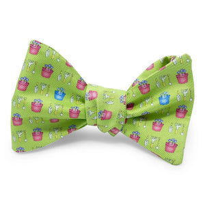 Introducing the BIRD DOG BAY PETER-BLAIR Garden Silk Bow Tie in Green, featuring a charming print of small pink and blue flower pots and white gardening tools. Handmade in North Carolina, this bow tie offers adjustable neck sizing for a perfect fit.
